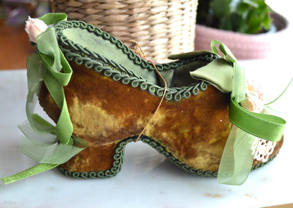 May include: A decorative shoe-shaped container in a vintage style. The shoe is made of brown velvet with green trim and ribbons. It features a high heel and is adorned with small flowers and lace.