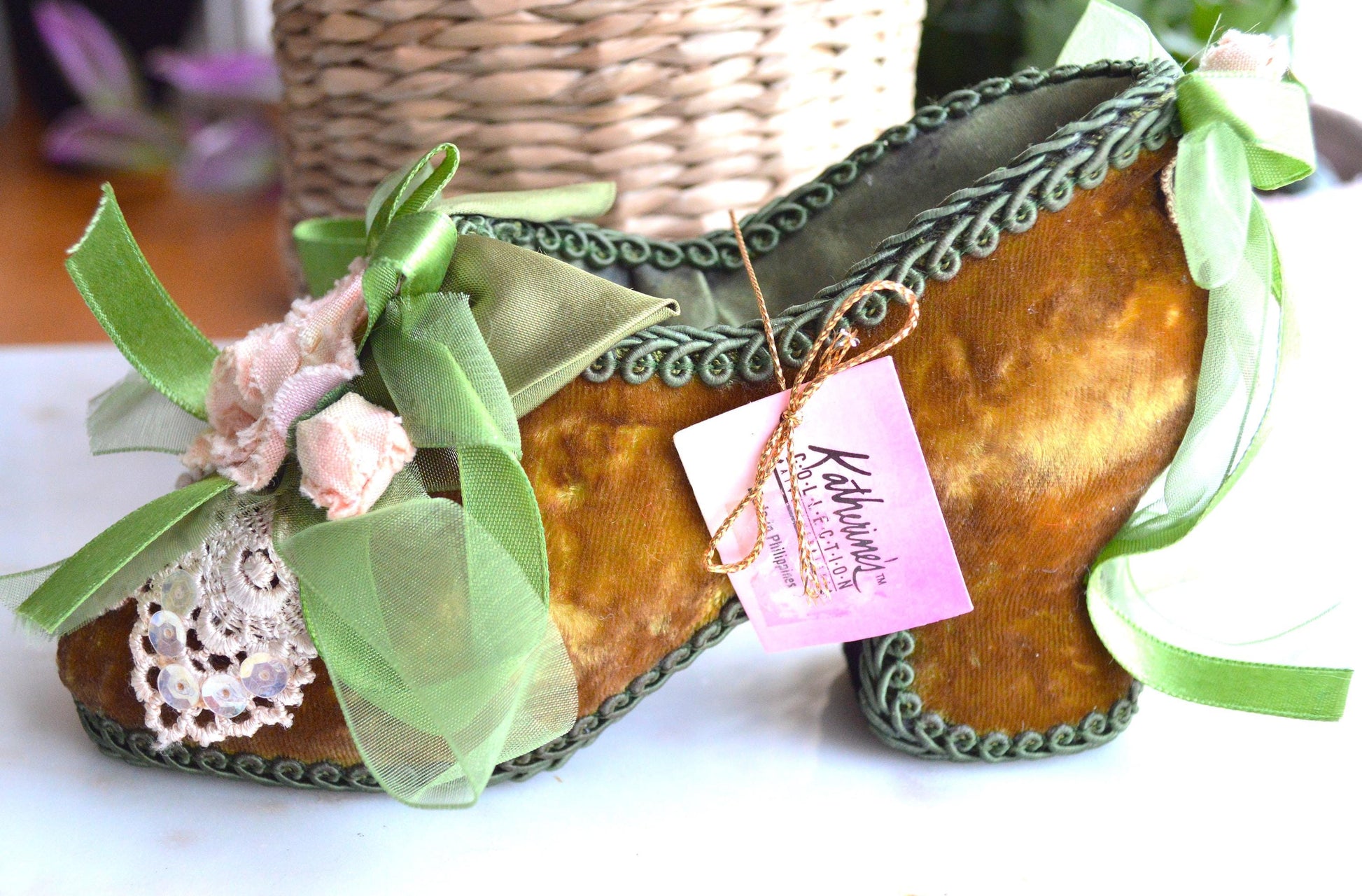 May include: A decorative, high-heeled shoe in a mustard yellow velvet material, adorned with green ribbons and lace. The shoe has a green trim and is embellished with small pink flowers. A pink tag with the text "Katherine's Collection" is attached.
