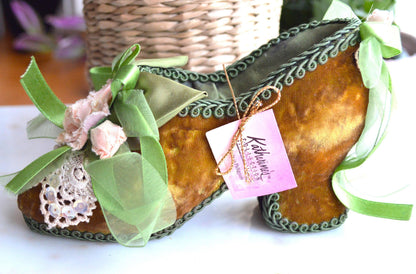 May include: A decorative, high-heeled shoe in a mustard yellow velvet material, adorned with green ribbons and lace. The shoe has a green trim and is embellished with small pink flowers. A pink tag with the text "Katherine's Collection" is attached.